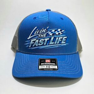 Blue and charcoal Richardson 112FP flat bill trucker hat featuring a white and blue "Livin' The Fast Life" racing graphic with checkered flag accents.