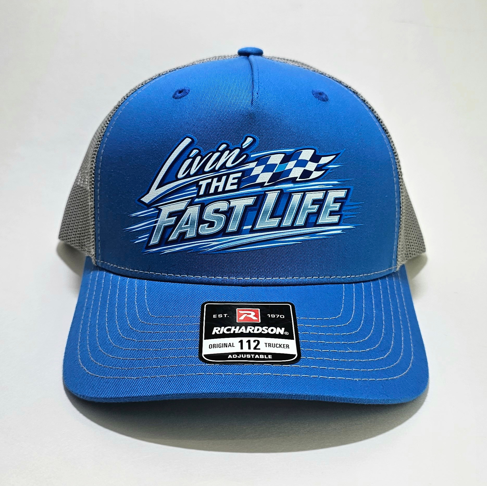 Blue and charcoal Richardson 112FP flat bill trucker hat featuring a white and blue "Livin' The Fast Life" racing graphic with checkered flag accents.